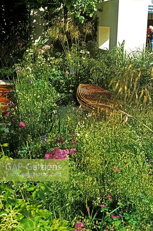 These Four Walls Garden at Chelsea Flower Show 2006 with bent wooden bench made from steamed Oak and Birch ply from New Zealand in woodland with planting of Stipa gigantea, Astrantia and Thalictrum - Thick wall with window in background 