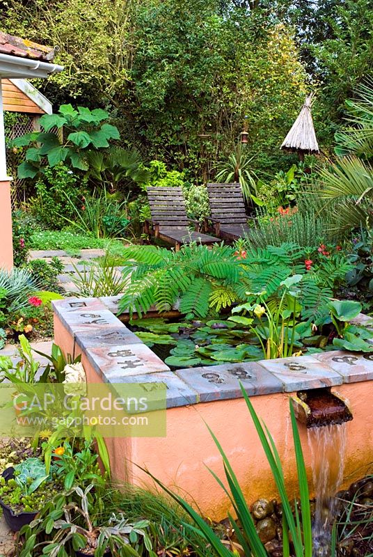 Raised terracotta painted pool water feature with cascade on terrace of exotic style garden with seating and tropical plants
