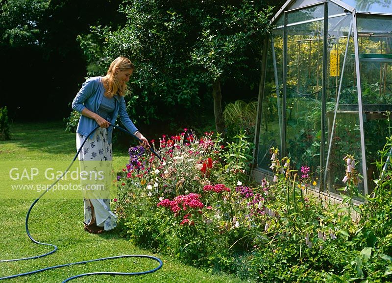 Woman watering flower bed with hosepipe next to greenhouse