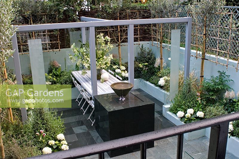 Grey steel painted frame supporting marble 'floating' table. Courtyard - 'enteraining' garden in whites, purple and green. Trellis with Olive trees trained in fan shape for privacy