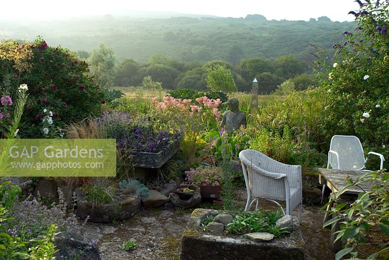 Seating area with alping sinks, self seeded plants, Rosa 'Roserie de L'Hay' and bog garden with statue and obelisk. View over Dinas Mountain and Garn Fawr peak in distance