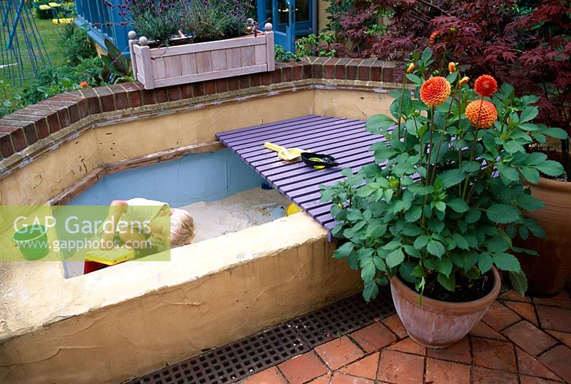 Patio with  young boy playing in sandpit made from breeze blocks and rendered with plaster. Softwood wooden decking covers the sandpit when not in use. Dahlias in terracotta pots.