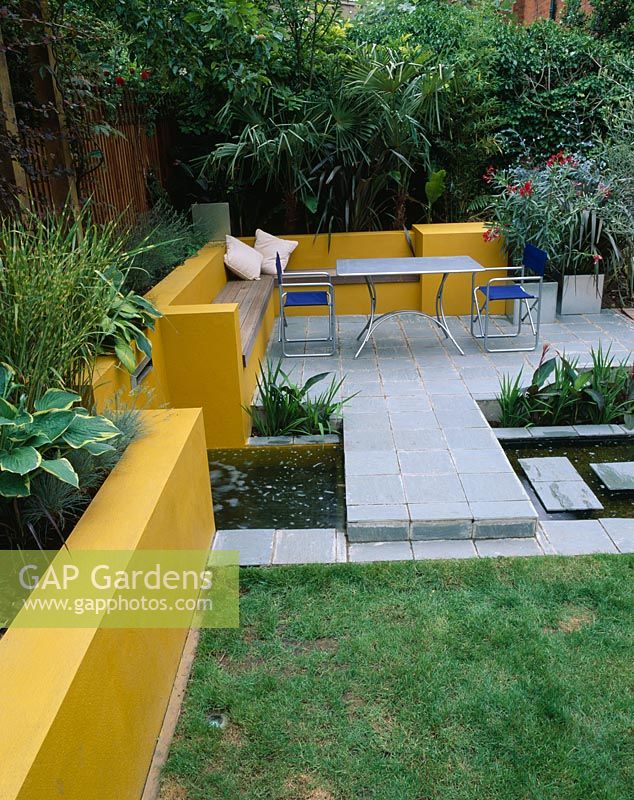 Aluminium table and chairs on patio surrounded by yellow rendered walls with raised beds and rill. Oleander in galvanised container. 