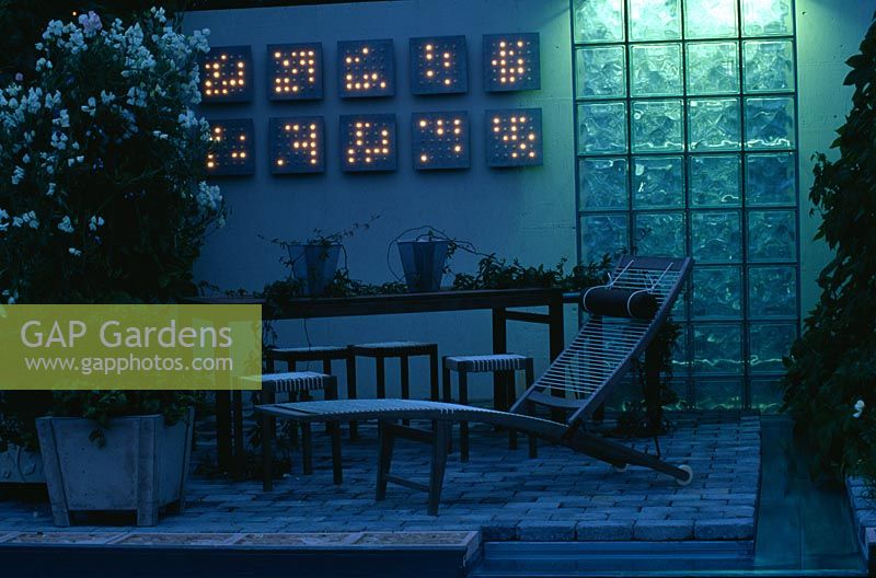 Modern balcony garden at night with wooden sunlounger, fairy lighting and glass screen water cascade and white Lathyrus odoratus - sweet peas in pot