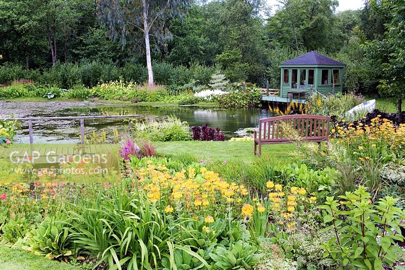 Acid bog garden. Stream with Astilbe arendsii, Astilbe 'Deutschland', Candelabra Primula. View to pond, bench and garden house.