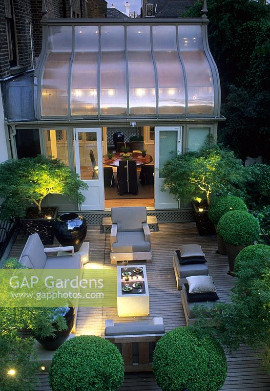 Overview of modern roof garden at night with lighting, table and chairs inside glass topped room and another outside. Containers of Buxus topiary spheres and Acers - Suleyman Garden, London 