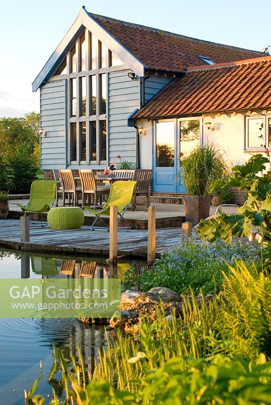 View across swimming pond to timber decking, outside eating area with table and chairs and converted barn with warm summer sunlight reflected on building - Carpe Diem, Bressingham, Norfolk