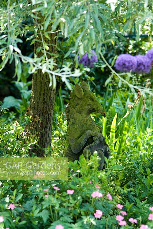 Stone statue of a Griffin like creature under a Pyrus salicifolia 'Pendula' - Weeping Silver pear tree, with a hardy Geranium in the foreground and Allium 'Globemaster' in the background.