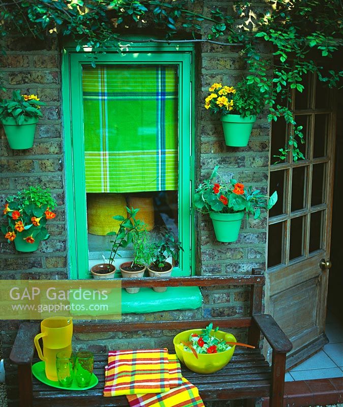 Colour co-ordinated containers hung on wall either side of window with green painted frame. Bowl of salad and jug and glasses on bench in front.