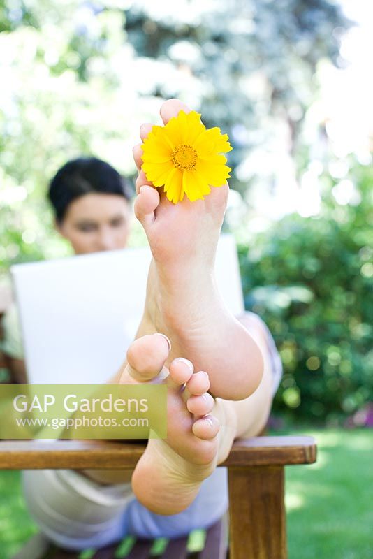 Woman sitting using laptop computer on wooden bench in the garden with yellow flower between her toes