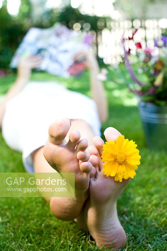 Woman lying on lawn looking at magazine with yellow flower between her toes