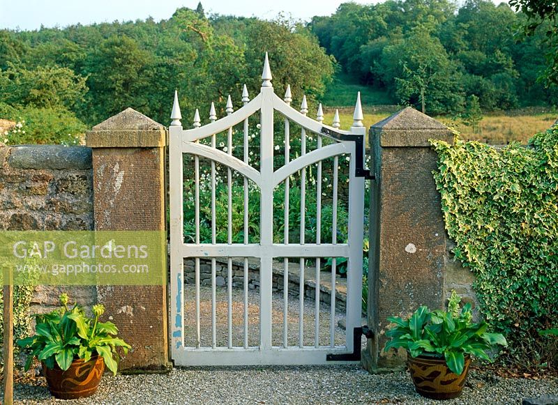 Wooden painted gothic style gate set between stone pillars and oriental pots with Eucomis and countryside beyond - Lawkland Hall, Yorkshire