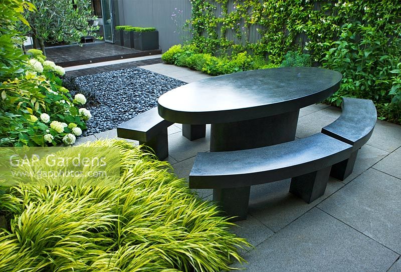 Small contemporary garden with black marble table and benches with Hakonechloa macra 'Alboaurea', Hydrangea and polished grey pebbles - London