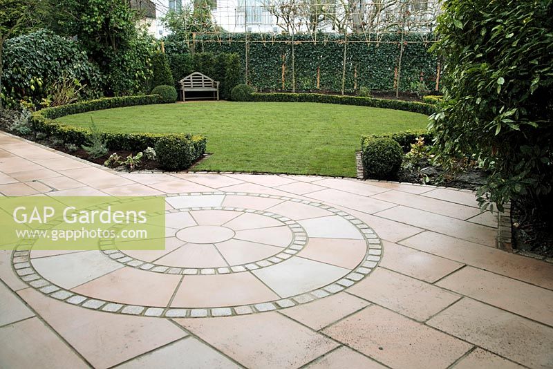 Circular patio design in Indian sandstone - cut and sandblasted, newly laid lawn in December, evergreen structural planting, curved Buxus hedge and pleached Carpinus betulus in London garden