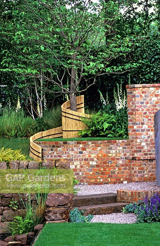 Garden with mix of hardscaping including brick wall, gravel, stone steps, drystone wall and natural reed screen. Digitalis in woodland planting - The Merrill Lynch Garden at RHS Chelsea Flower Show 