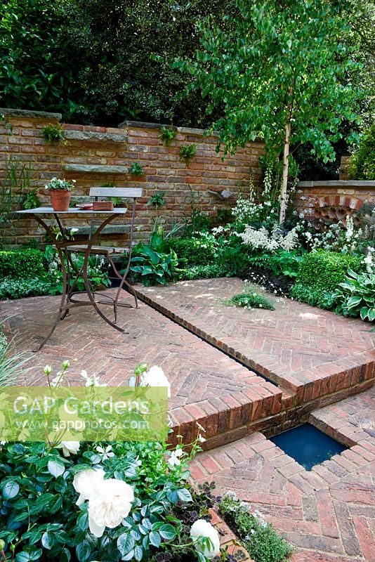 Table and chair with pot of Viola 'Wisley White'on brick-weave terrace with rill and small pool. Betula utilis var. jacquemontii in corner with white and green underplanting including Lamprocapnos spectabilis 'Alba', white Astilbe, white foxgloves Digitalis purpurea f. albiflora and white Polemonium. Rosa 'Macmillan Nurse' in foreground, Erigeron karvinskianus in crack in paving. Jacob's Ladder Garden, sponsored by Hewitt Landscapes Ltd - Silver Flora medal winner for Courtyard Garden at RHS Chelsea Flower Show 2009