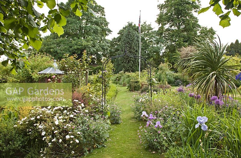 Overall garden view of mature shrubs and trees, archways, grass paths and summerhouse - Millennium Garden NGS, Lichfield, Staffordshire
