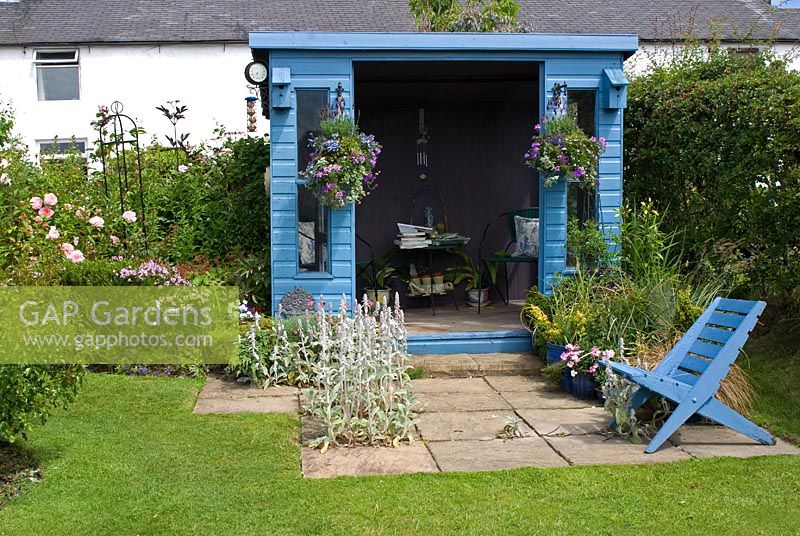 Blue painted wooden summerhouse with hanging baskets, and pots with blue seat on patio in cottage garden at New Row Cottages, NGS garden, Lancashire