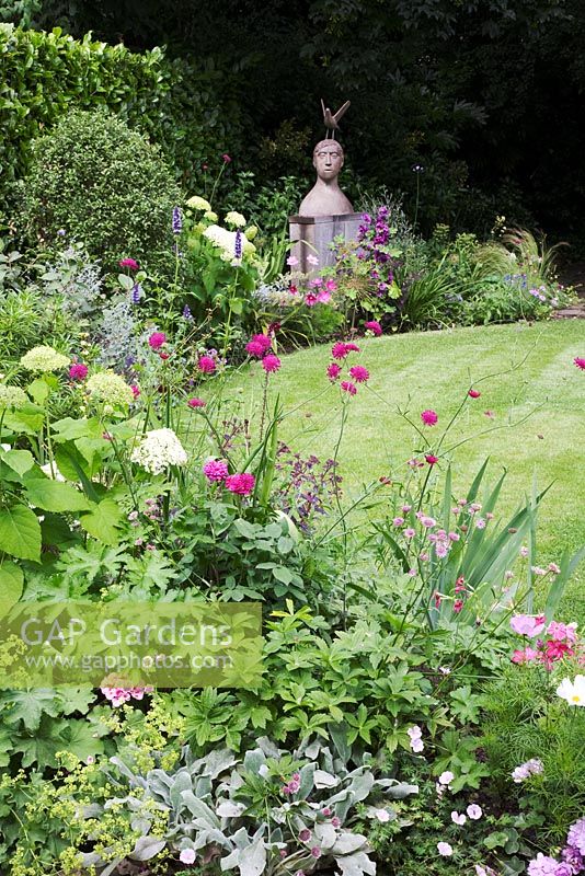 Small Urban garden packed full of plants simply designed around a central circular lawn. Flower border surrounding circular lawn with focal point sculpture by Christopher Marvell, Hydrangea arborescens, Knautia macedonica, pink Astrantias, and Iris foliage.
