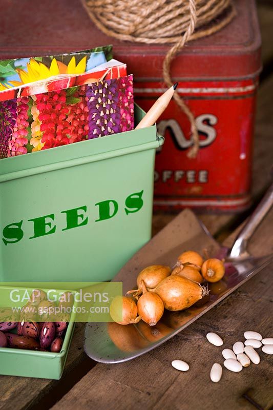 Fairtrade seed tin with seed packets, garden string, Runner bean seed 'Prizewinner stringless', stainless steel trowel with Onion sets on an upturned wooden crate
 