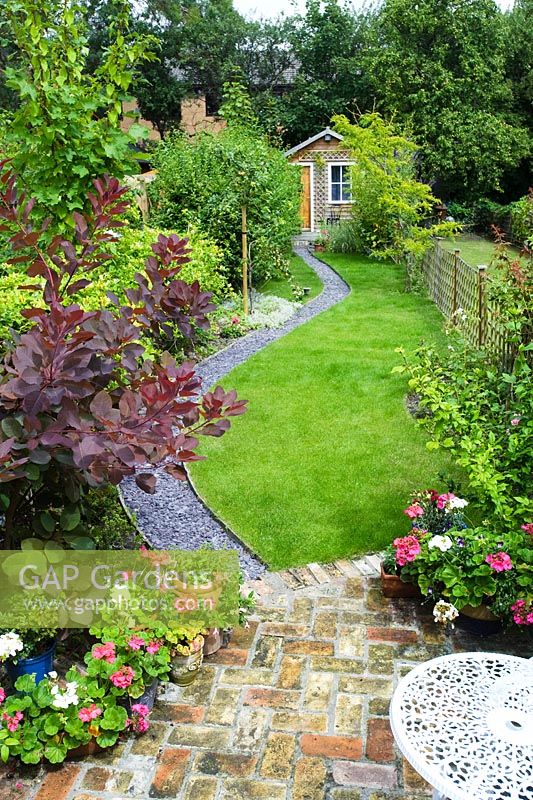 Elevated view of long, narrow town garden with lawn and sinuous path leading to garden studio. Brick paving, slate chippings, Cotinus, pots of Pelargoniums.