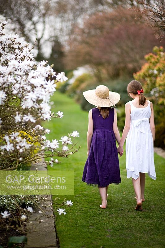 Two girls wearing summer dresses walking along a grass path between borders of shrubs and Magnolia