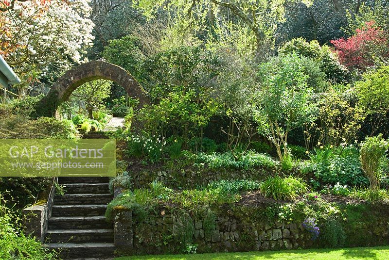 Circular brick entrance into the kitchen garden at the top of steps leading up from the lawn, with magnolia flowering beyond - Greencombe Garden, Porlock, Somerset