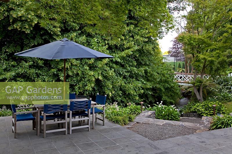 Suburban garden with seating area next to stream. A large Tilia - Lime tree on a neighbouring property across the stream forms a green screen. Christchurch, New Zealand