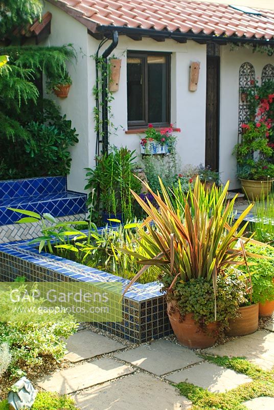 Paved courtyard with raised pool and seat clad in blue glazed tiles. Phormium in container.