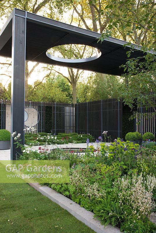 A louvered timber cloister above a circular pond. Plants including Geranium phaeum, Iris sibirica 'Tropic Night', Euphorbia, Anthriscus sylvestris 'Ravenswing' -Queen Anne's Lace and Dicentra formosa. The Cancer Research UK Garden, Gold Medal Winner RHS Chelsea Flower Show 2010
 