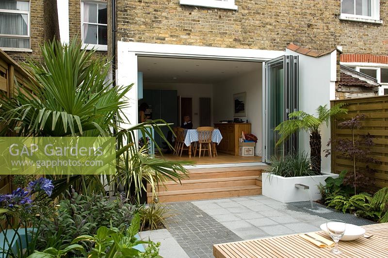 Small urban garden with with paved patio area and sliding French door, Trachycarpus to left and small water feature with spout