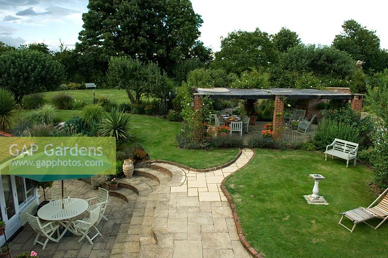 Elevated view of large country garden with lawn, pergola and dining area on terrace with curved steps. Parsons Cottage, Essex, UK