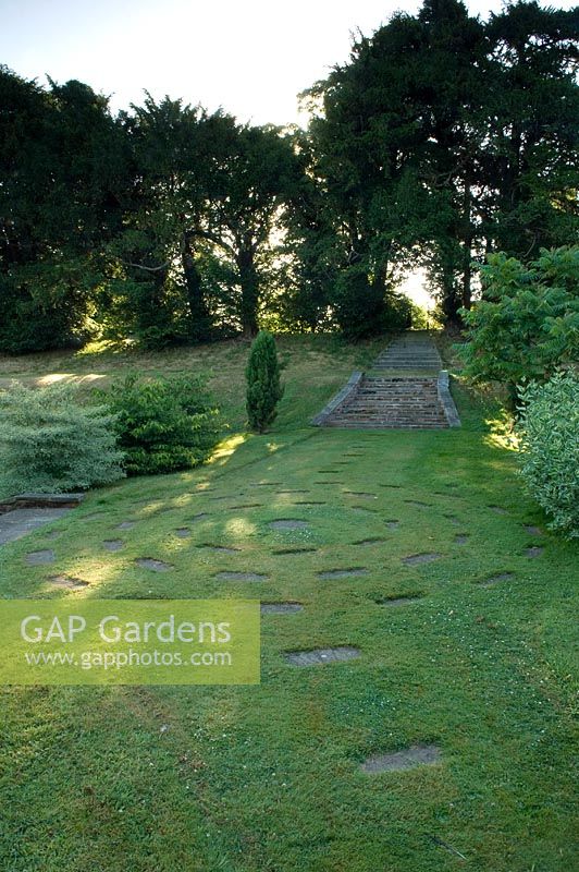 Round lawn with circle of paving slabs and steps up to mature trees. Robinson garden, Ousden House, Suffolk