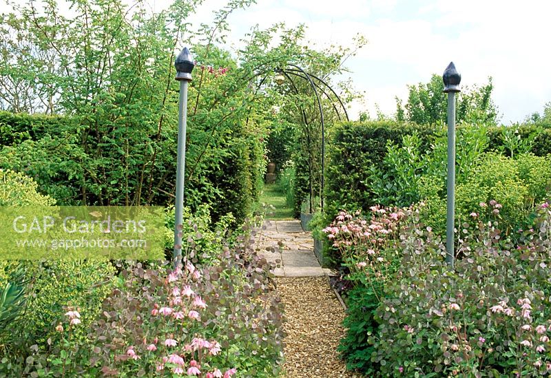 Path leading through finial topped posts, framing the view to metal arbour and pot beyond. Fovant Hut Garden, Wilts