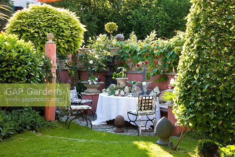 Iron garden chairs with a display of cones, vases and urns on the table, surrounded of a wall, a plinth and formal clipped shrubs - containing planting of Carpinus betulus, Catalpa bignonioides 'Aurea', Prunus laurocerasus and Vitis