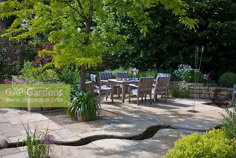 Raised dry stone wall bed, a rest area with wooden furniture and a meandering canal through the flagstone paving. Planting includes Agapanthus, Allium christophii, Centranthus ruber 'Coccineus', Gleditsia triacanthos 'Sunburst' and Tanacetum parthenium 