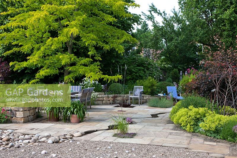 Flagstone paved area with meandering canal leading to a pebble beach zone, a Gleditsia makes a focal point and gives light shade to garden furniture. The background is formed by trees and a curved dry stone wall. Planting includes Alchemilla mollis, Allium christophii and Thymian