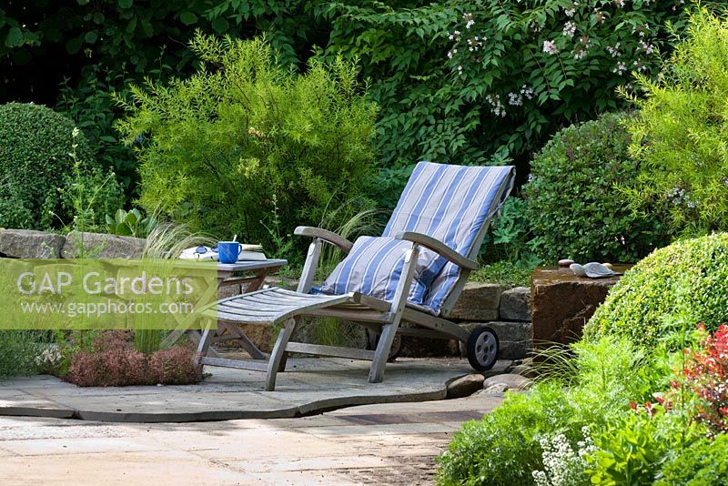 A wooden deck chair with blue striped cushions, dry stone wall and a meandering canal that runs through flagstone paving. Planting includes Allium christophii, Buxus, Kolkwitzia amabilis, Sedum album, Spiraea thunbergii and Stipa pennata