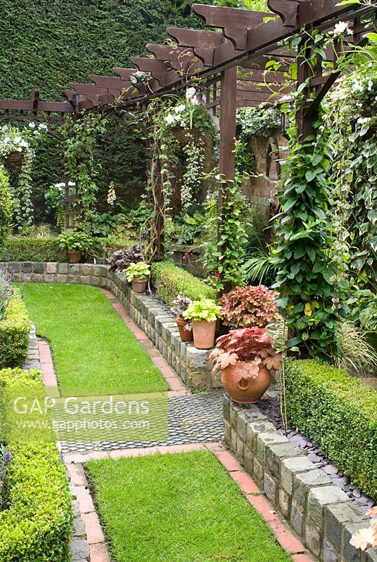 Secluded part of garden with paths and walls made of small reclaimed cobbles, station platform tiles and bricks, Buxus sempervirens hedges, potted Heuchera and trellis with clematis and hanging baskets - Brocklebank Road, Southport, Lancashire NGS 
