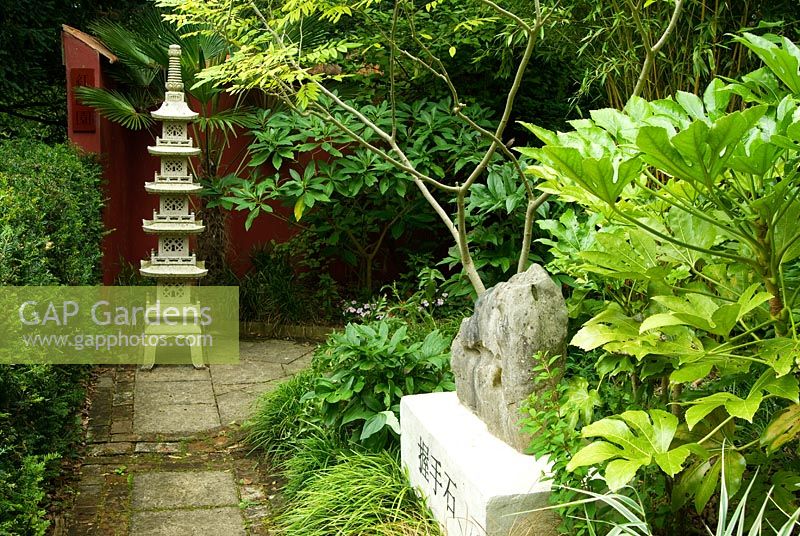 Chinese granite pagoda in the Red Wall garden planted around with palm Trachycarpus wagnerianus and Edgeworthia chrysantha. 'Holding hands stone' on plinth in foreground surrounded by Bamboo, Fatsia japonica and Cladrastis sinensis. Beggars Knoll, Newtown, Westbury, Wiltshire, UK