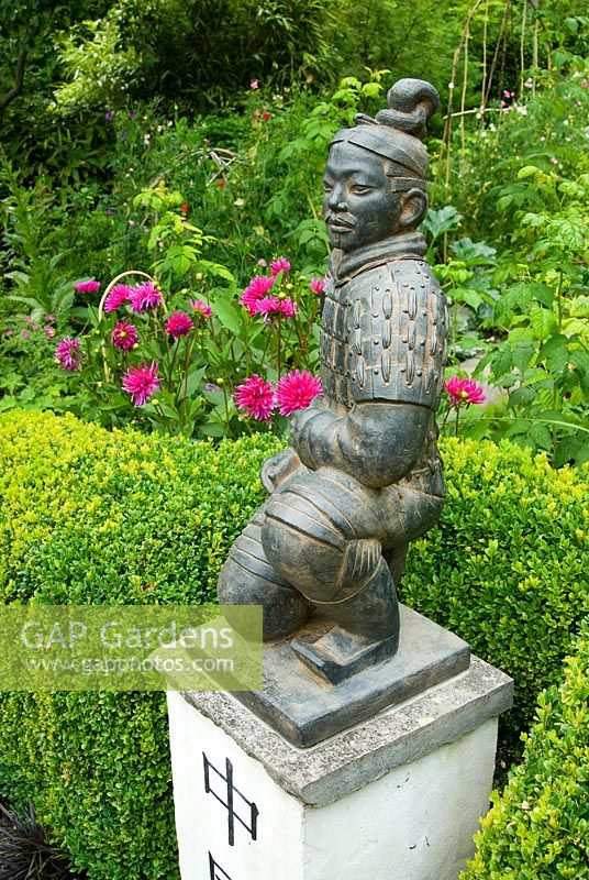 Statue of a terracotta warrior from Xi'an crouching on a pedestal marked with the word 'China', framed by clipped Buxus - Box hedge and Dahlias. Beggars Knoll, Newtown, Westbury, Wiltshire, UK