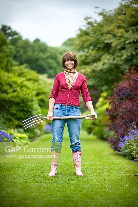 Woman wearing blue jeans and wellies standing on a lawned path carrying a garden fork 