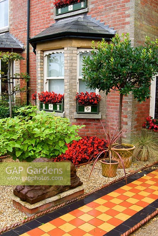Front garden of Victorian terraced house features a tiled path beside gravelled area containing statuary and planting including Begonia 'Big Boy' and Cordylines. The Secret Garden at Serles House, Wimborne, Dorset, UK