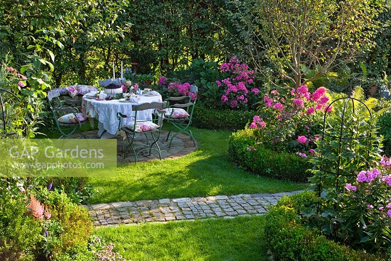 Round paved seating area with dressed table and wooden iron chairs next to planting with Buxus and Phlox paniculata