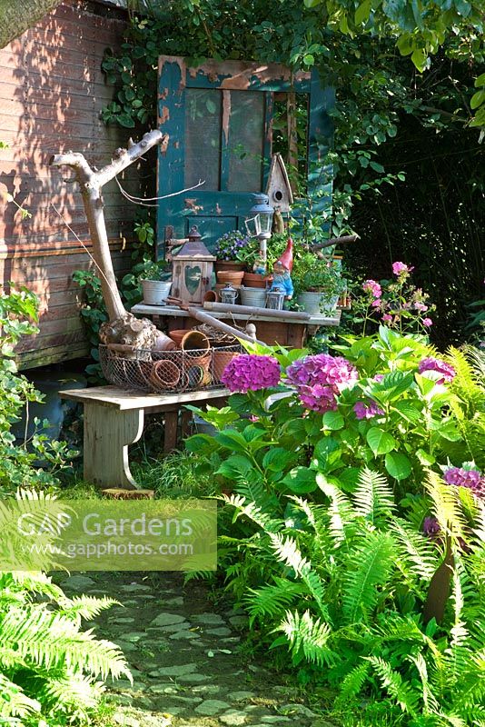 Fanciful collection of garden objects with pots, garden gnomes and bird boxes next to, Hydrangea macrophylla and Matteucia struthiopteris