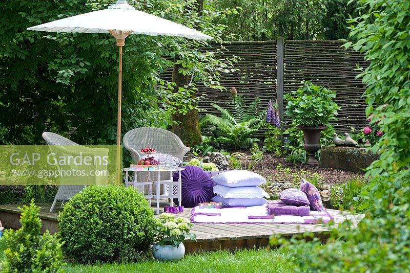 Wooden deck with white wicker garden furniture, parasol and a blanket with white and lilac cushions. Plants are Buxus, Fern, Hydrangea macrophylla and Lupinus