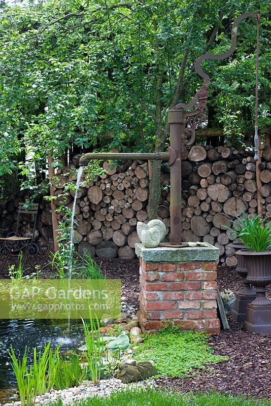 Water well next to pond, with two stone sculptured snails, metal urns and a wood pile in the background. Plants include Lysimachia nummularia, Salix and Typha 