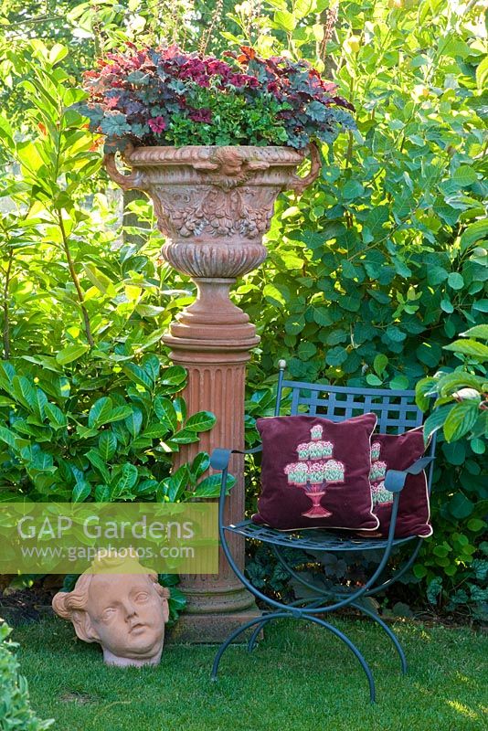 Terracotta urn on a plinth next to metal chair with cushions and terracotta head. Plants are Cotinus coggygria, Heuchera micrantha 'Palace Purple', Pelargonium and Prunus laurocerasus 