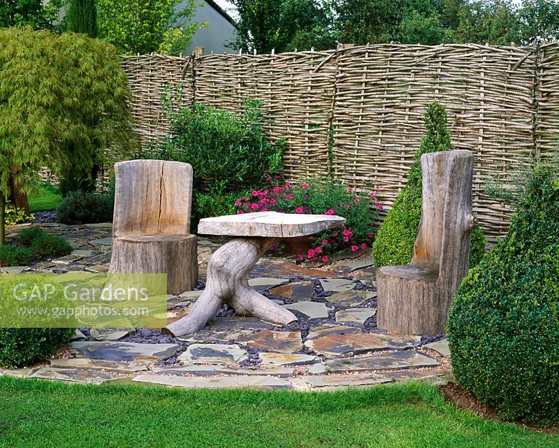 Wooden furniture on slate terrace with box topiary shapes and willow fence 