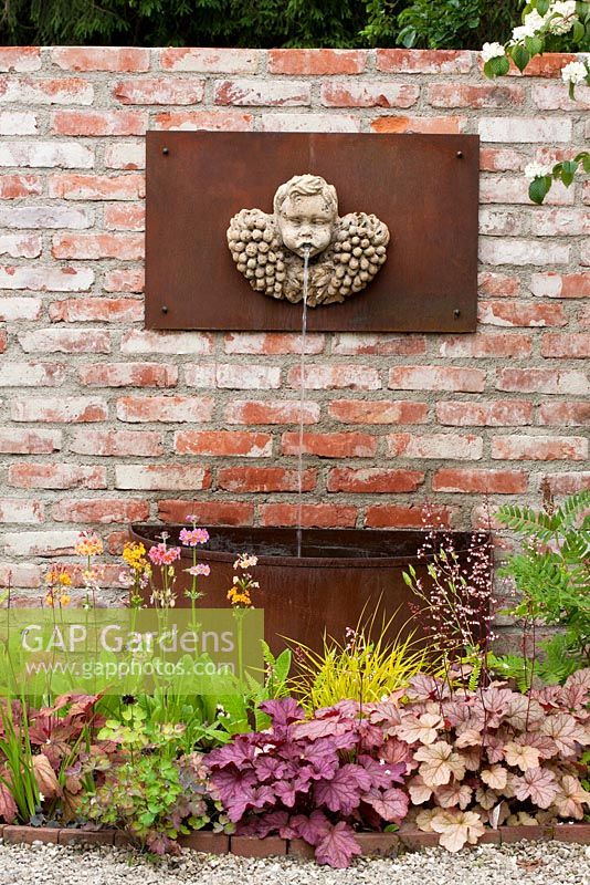 The colours of a putto fountain with rusty basin on a brick  wall are reflected in a planting of Primula bullesiana (P beesiana x bulleriana)